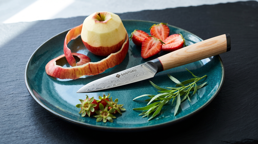 A Damascus-clad paring knife on a teal ceramic plate with a half-peeled apple and strawberries